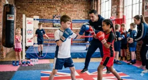 A male coach instructing a young boy and girl during a sparring session in a traditional UK boxing gym.