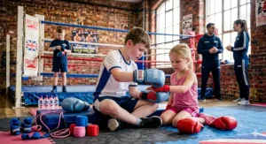 An older boy helping a young girl fit her blue junior boxing glove over red hand wraps while sitting on gym mats.
